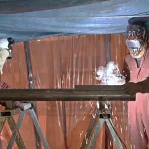 Two workers in protective gear weld a metal pipe in a screened area with sparks