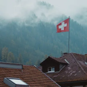 Swiss Flag on alpine rooftops, mouintain in the background with rolling clouds from Emission Impossible film by Floating Harbour