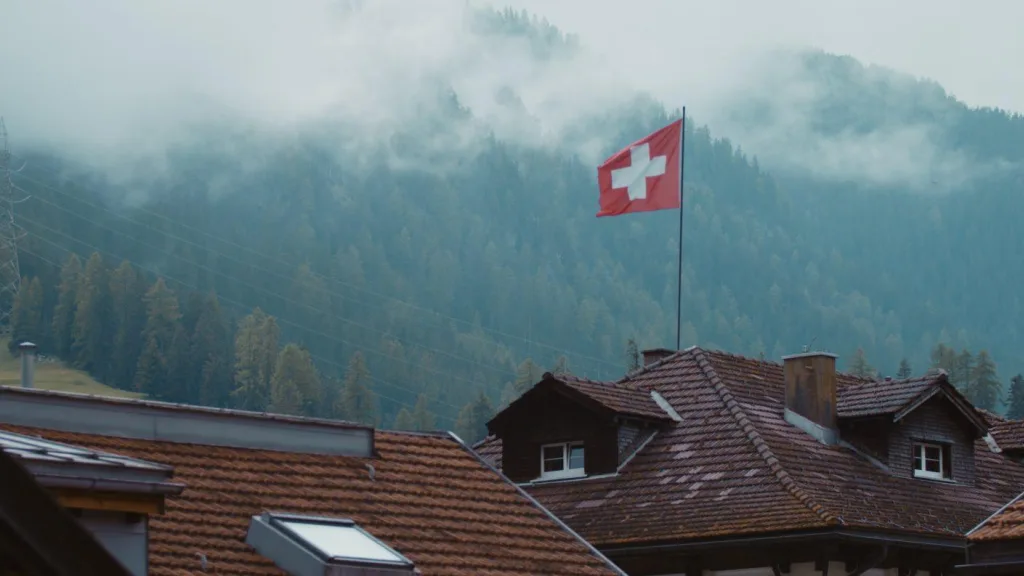 Swiss Flag on alpine rooftops, mouintain in the background with rolling clouds from Emission Impossible film by Floating Harbour