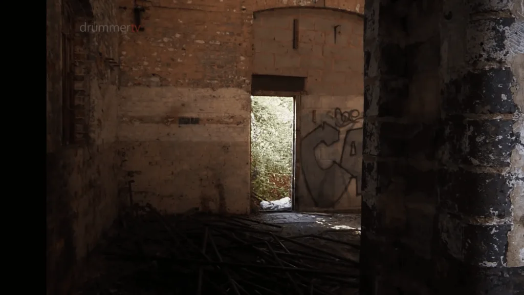 An image of a derelict building interior with crumbling brick walls, scattered metal pipes on the floor, and a doorway leading to sunlit foliage, used in the location filming for the BBC documentary Where am I sleeping tonight?