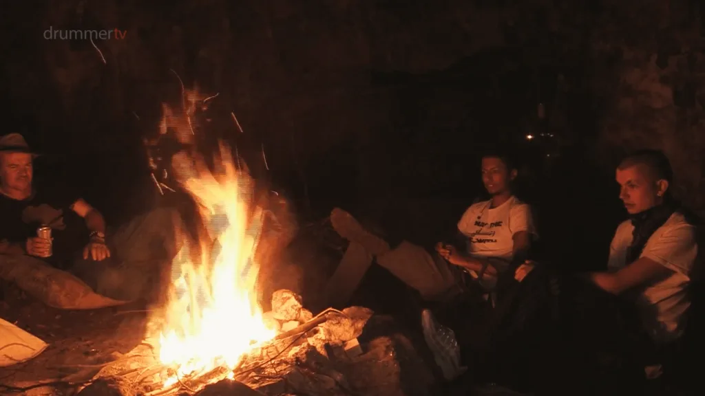 Three people sit around a large, bright campfire inside a cave during location filming for the BBC and Drummer TV documentary series, Circus Kids.