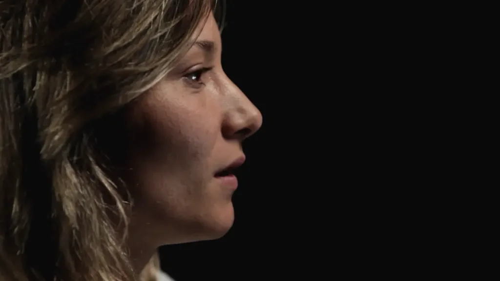 Close-up profile of a woman speaking on camera against a dark background.