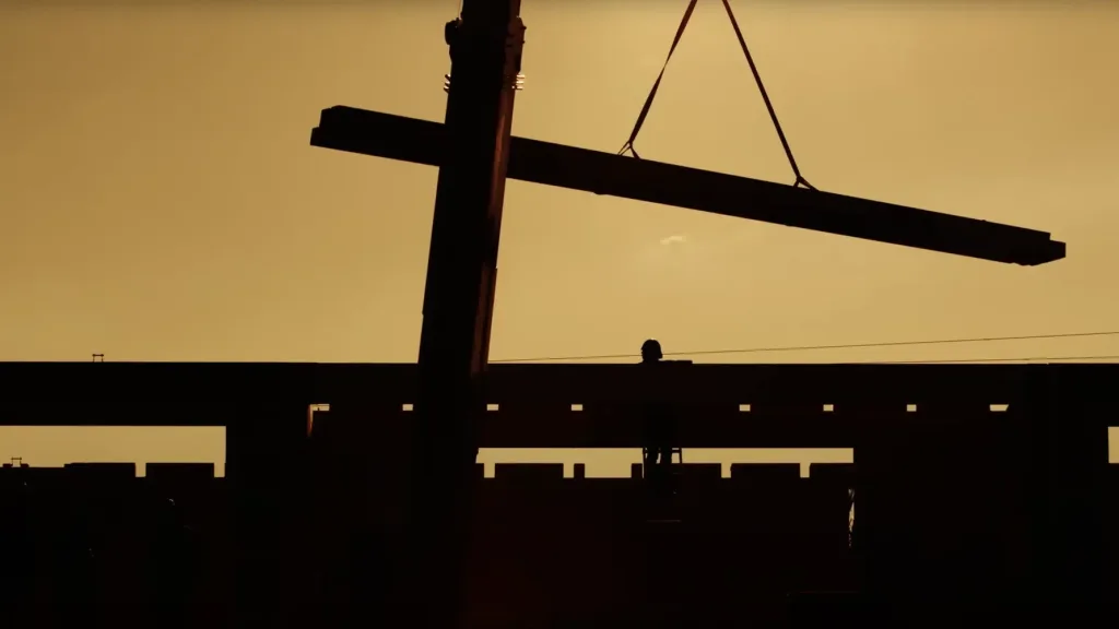 Silhouette of a construction site with a large beam being lifted by a crane at sunset.