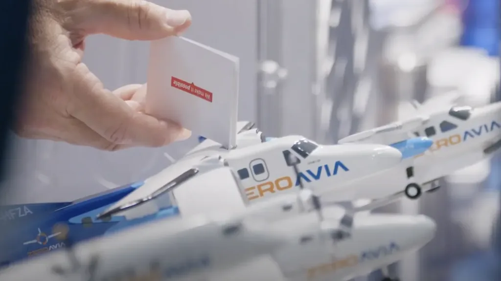 Hand holding a small business card over a miniature ZeroAvia aircraft model at an industry exhibition stand filmed by Floating Harbour.