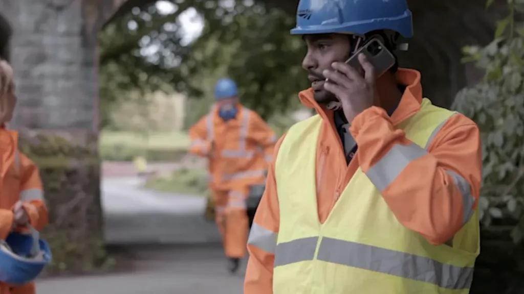 Railway workers in high-visibility clothing and helmets stand under a bridge while one talks on a phone
