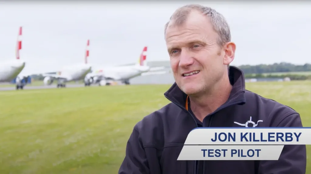 Test pilot speaking during an interview at an airfield with aircraft in the background.