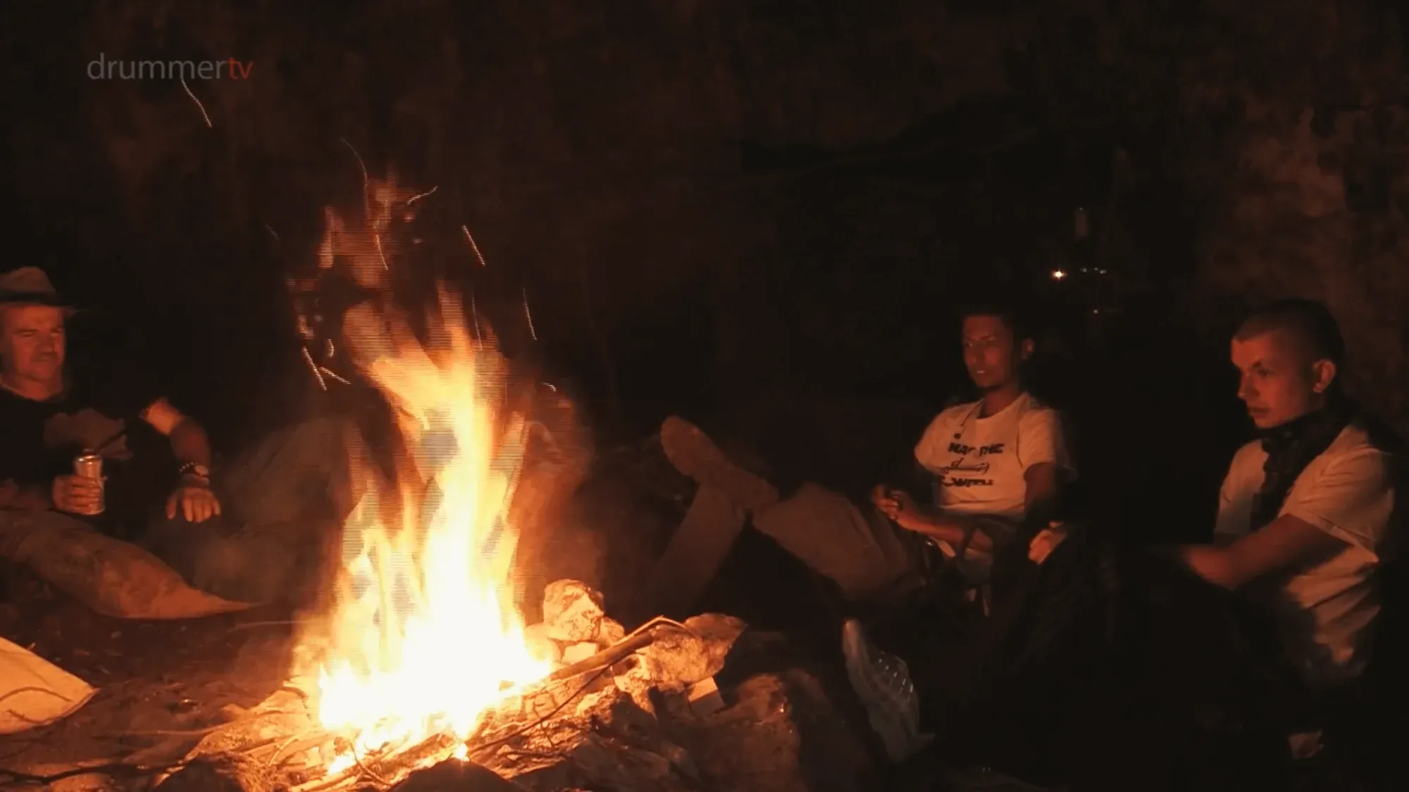 Three people sit around a large, bright campfire inside a cave during location filming for the BBC and Drummer TV documentary series, Circus Kids.