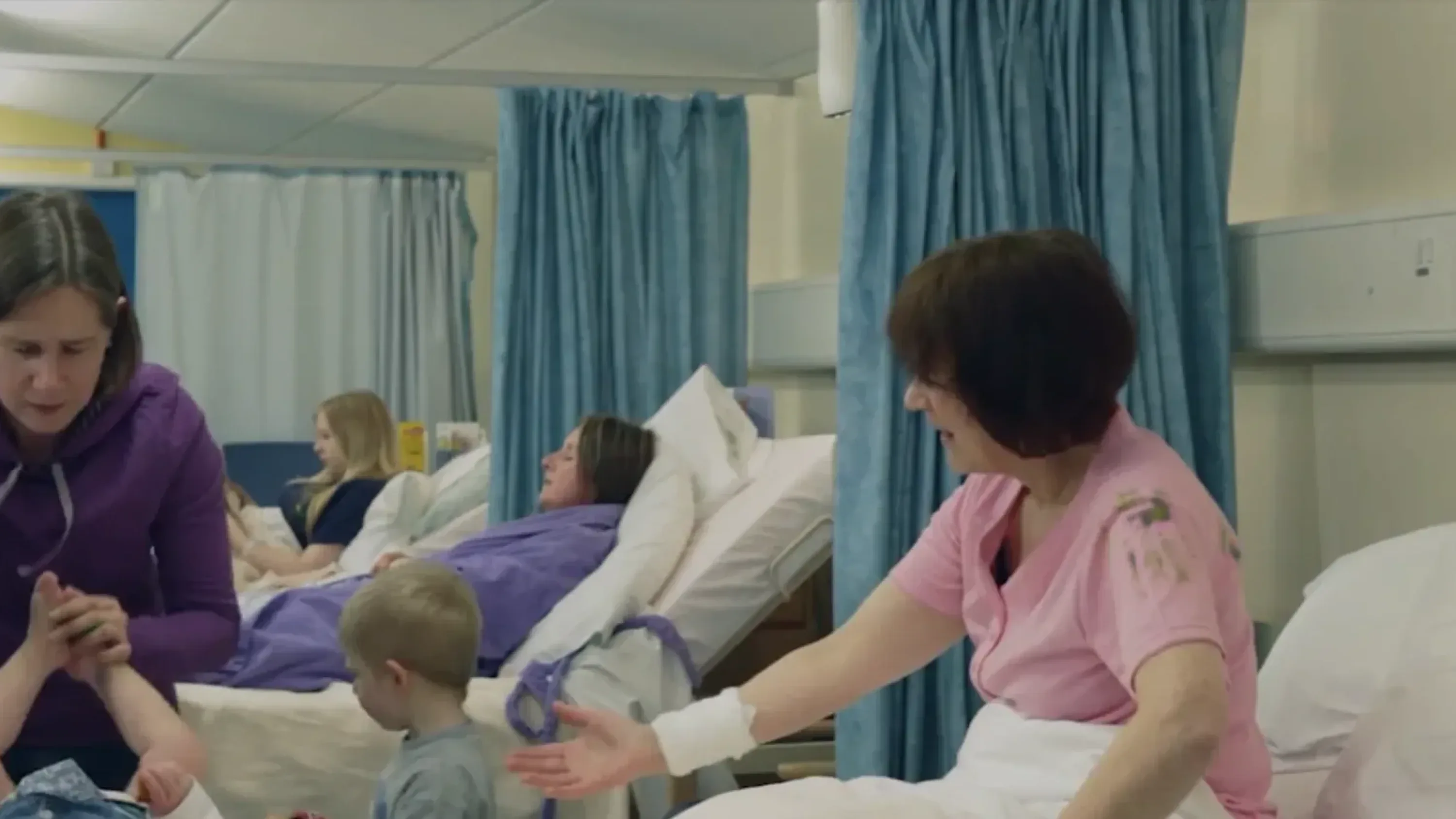 Patients and carers interact on a hospital ward with blue privacy curtains and medical beds