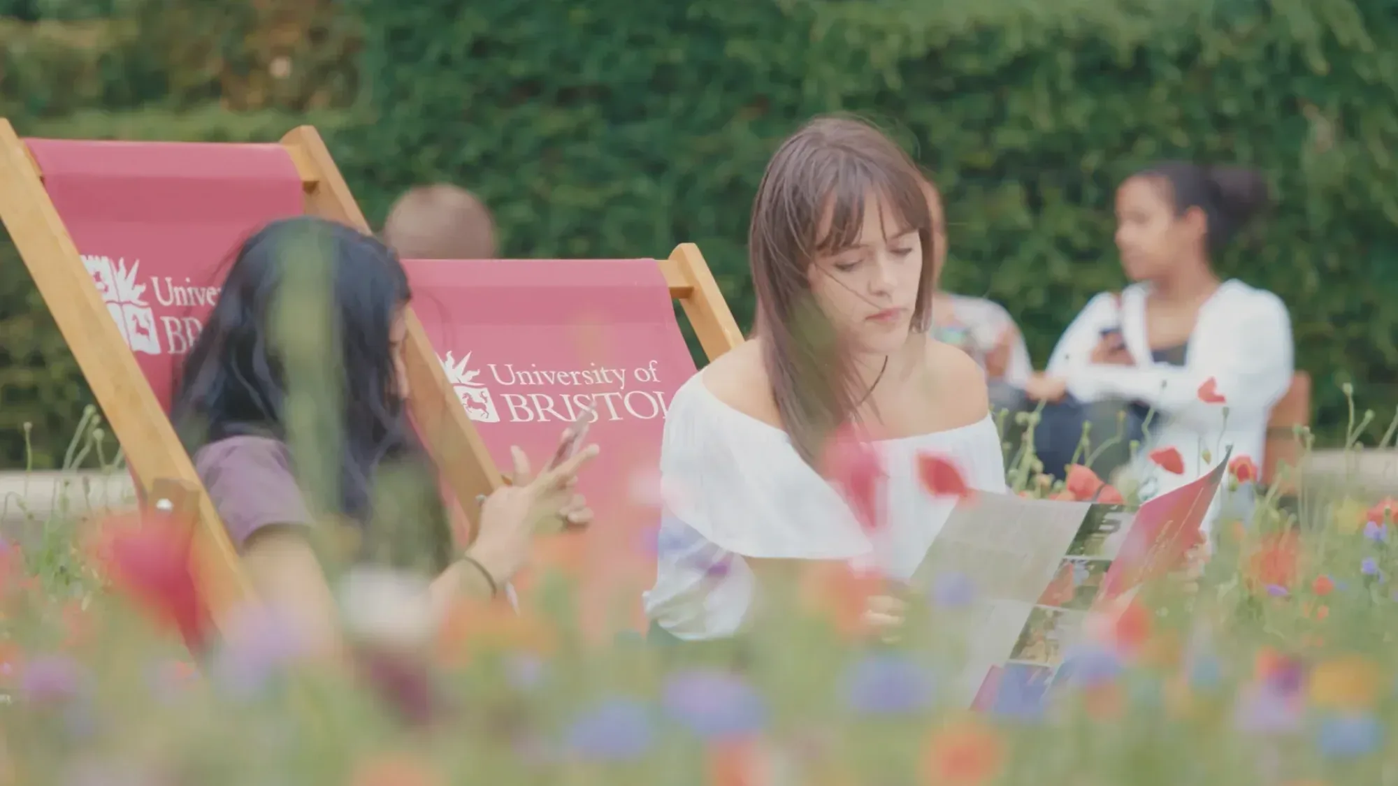 Students sit on University of Bristol deckchairs in a garden reading and chatting amongst flowers
